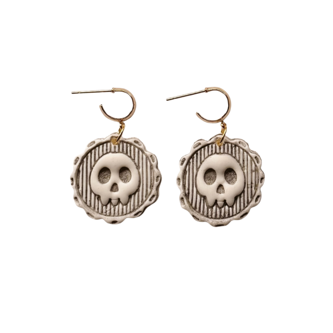 A pair of gothic-style drop earrings featuring circular silver-toned medallions with a raised skull motif and vertical textured stripes, suspended from gold-toned fishhook wires, shown against a plain white background.