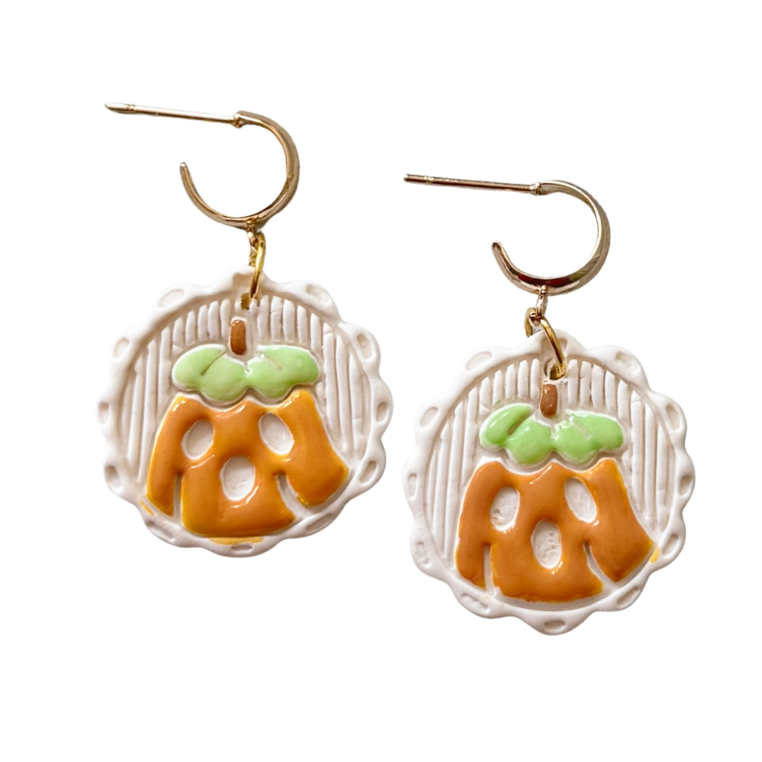 A pair of dangling earrings with gold-tone huggies and round white clay pumpkin charms featuring orange faces and light green stems, shown on a plain white background.